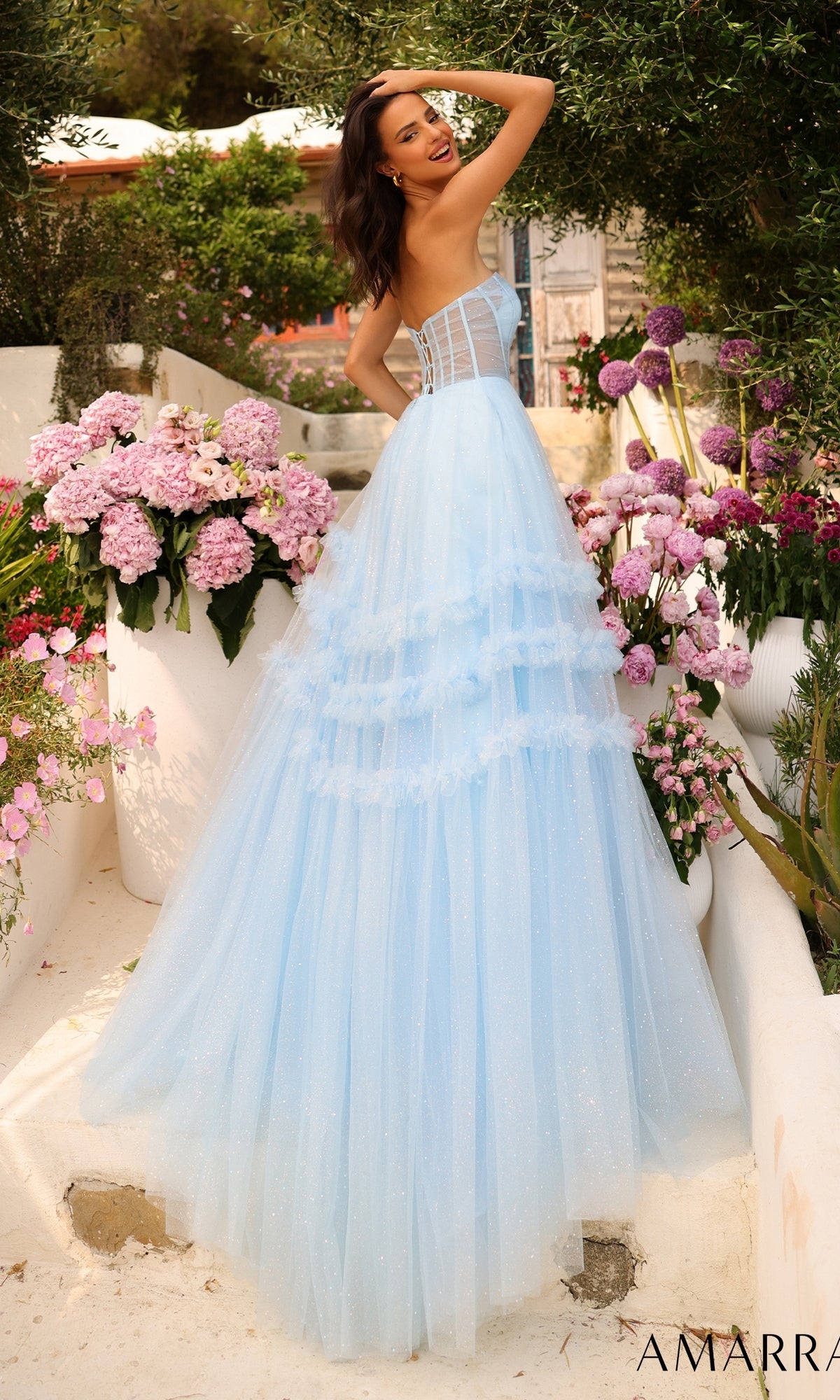 Woman wears the Amarra Strapless Sheer-Bodice Prom Ball Gown 88794, featuring a glitter-tulle overlay, posing outdoors among large vases of pink and purple flowers with one hand on her head.